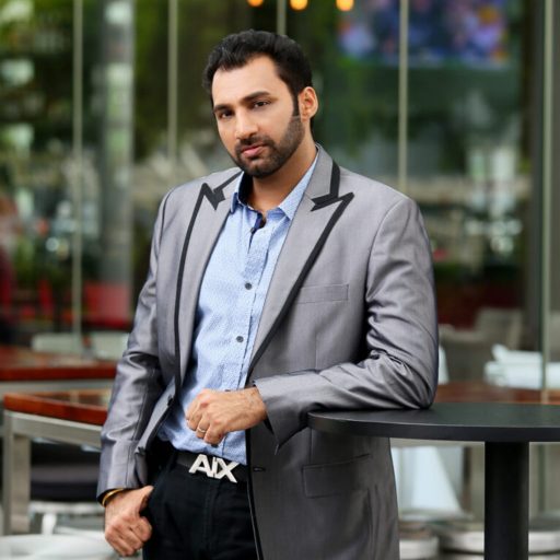 Roger Khera – Singapore Property Agent and DJ Roger khera in a grey blazer posing confidently at a modern outdoor café with stylish lighting and glass backdrop.
